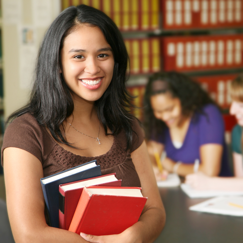 Young Hispanic College Student Holding a Stack of Colorful Books in Campus Library with People Behind Her.